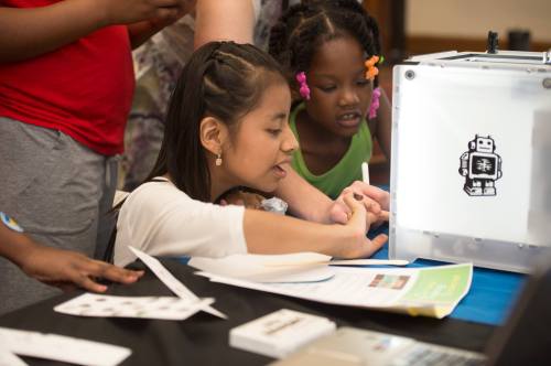 A girl focuses on a project at a table, while another child looks on, surrounded by papers and a lightbox.