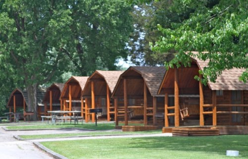 Row of wooden cabins with pitched roofs, surrounded by green grass and trees in a park setting.