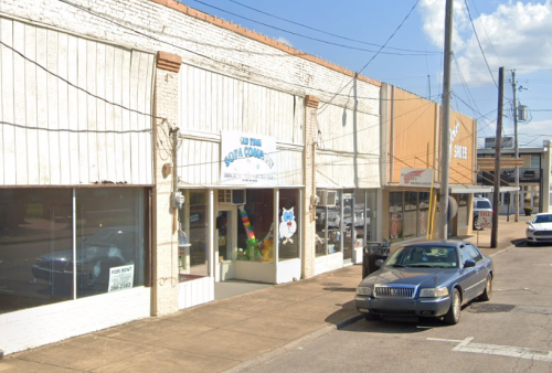 A street view of a small commercial area with shops, a parked car, and clear blue skies.