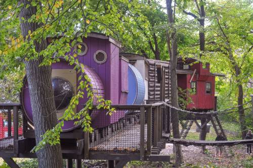 Colorful treehouse resembling a train, nestled among green trees, connected by a net bridge.