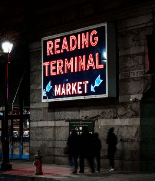 Neon sign for Reading Terminal Market illuminated at night, with people walking by and a streetlamp in the foreground.