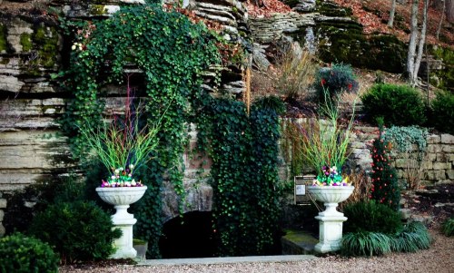 A stone wall covered in greenery, flanked by two decorative planters with colorful plants.