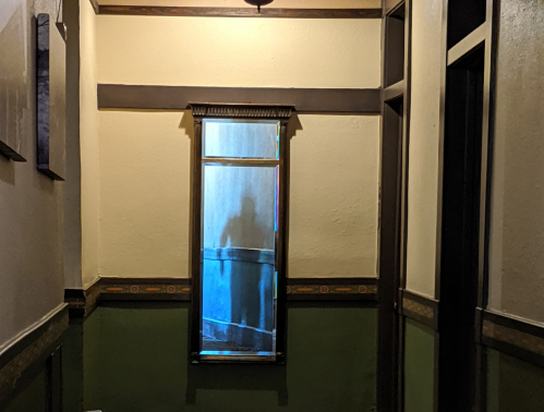 A dimly lit hallway with a mirror reflecting a shadowy figure and blue light at the end.