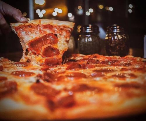 A hand lifts a slice of pepperoni pizza from a whole pizza, with seasoning jars in the background.