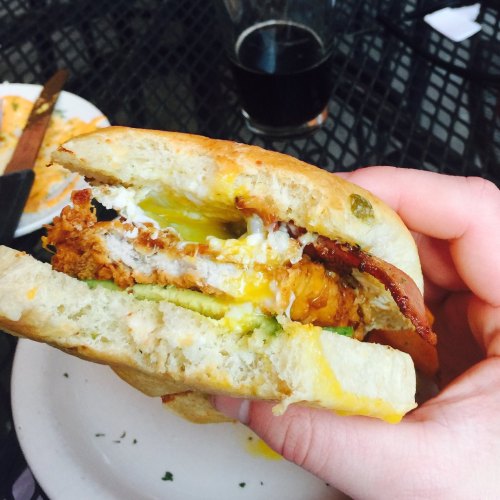 A hand holding a sandwich with layers of fried chicken, egg, and green toppings, with a drink in the background.