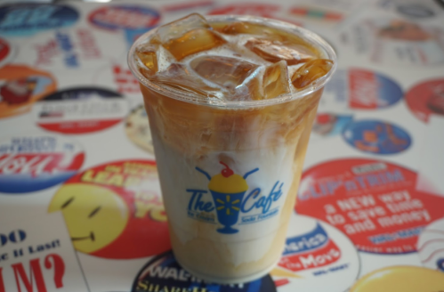 A clear cup filled with iced coffee and cream, sitting on a table covered in colorful stickers and logos.