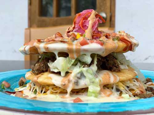 A towering plate of layered tacos with ground beef, lettuce, cheese, and drizzled sauces, garnished with pickled onions.