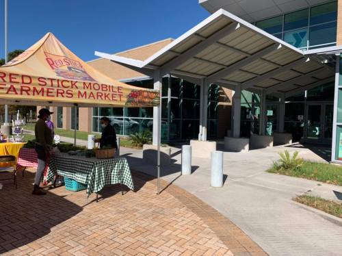 A farmers market stall with a canopy outside a modern building, featuring tables with produce and shoppers nearby.