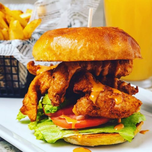 A crispy chicken sandwich with lettuce, tomato, and sauce, served with fries and a drink in the background.