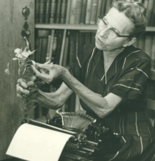 A woman in glasses arranges flowers while sitting next to a typewriter, surrounded by bookshelves.