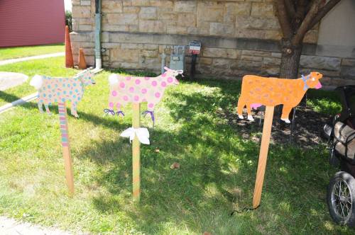 Three colorful, painted cow cutouts on wooden stakes, displayed on grass near a stone wall.