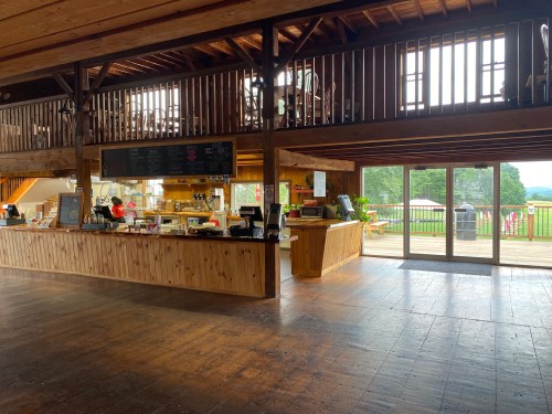 A rustic café interior with wooden beams, a counter, and large windows overlooking a green landscape.