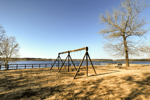 A swingset by a calm lake, surrounded by trees and a clear blue sky, with a sandy area in the foreground.