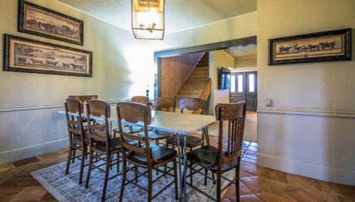 A dining area with a wooden table and chairs, framed artwork on the walls, and a view of a staircase.