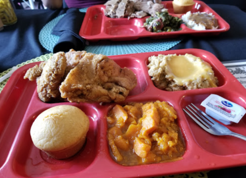 A red tray with fried chicken, cornbread, mashed potatoes, sweet potatoes, and a packet of sauce.