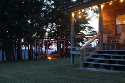 A cozy cabin at dusk, with string lights, a fire pit, and swings in a serene outdoor setting.