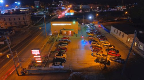 Aerial view of a parking lot at night, filled with cars, with city lights and buildings in the background.