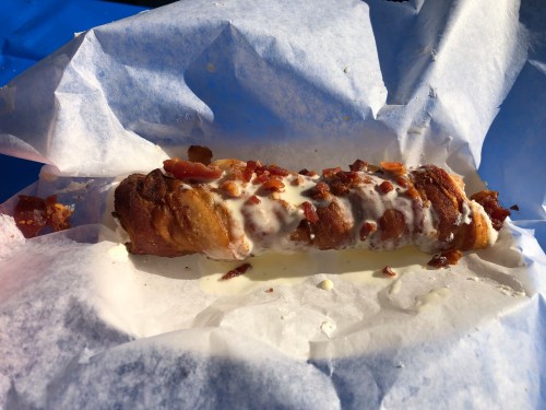 A crispy pastry filled with cream and topped with crumbled bacon, resting on white parchment paper.