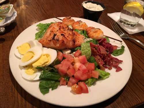 A plate of grilled salmon, shrimp skewers, salad with tomatoes, bacon, and sliced hard-boiled eggs, with a side of sauce.