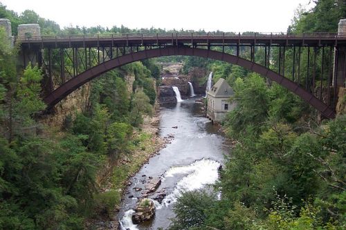 A metal bridge spans a river with waterfalls, surrounded by lush greenery and rocky terrain.
