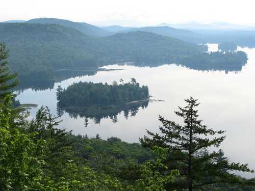 A serene landscape featuring a calm lake surrounded by lush green hills and distant mountains under a cloudy sky.