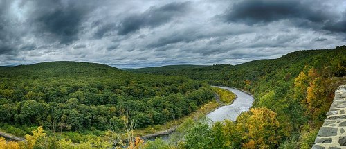 A panoramic view of a winding river surrounded by lush green hills under a cloudy sky. Autumn colors peek through the trees.