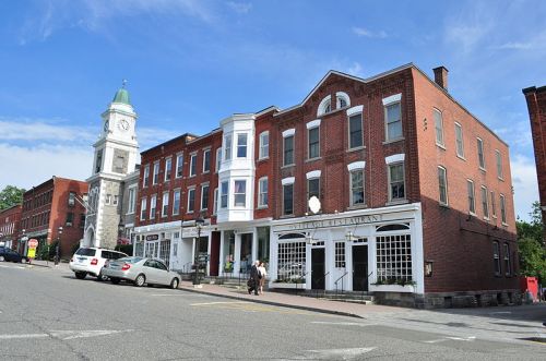 Historic brick buildings line a street, featuring a clock tower and a restaurant with outdoor seating.
