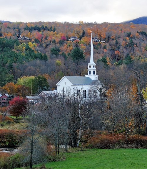 A white church with a tall steeple surrounded by colorful autumn trees and a serene landscape.