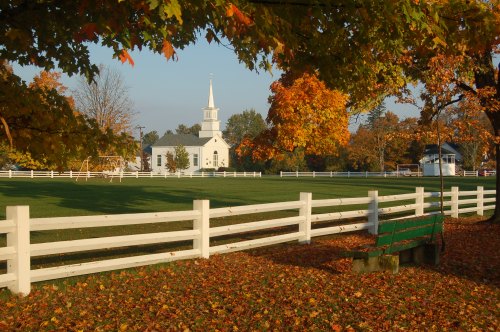 A white church with a steeple surrounded by colorful autumn trees and a white fence, with fallen leaves on the ground.