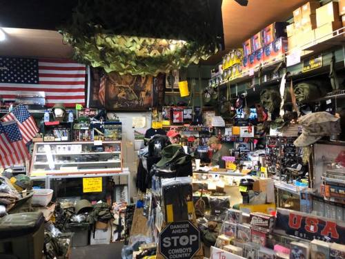 A cluttered military surplus store filled with gear, clothing, and decorations, including flags and camouflage netting.