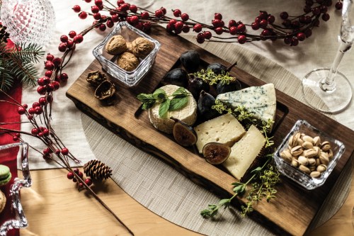 A wooden cheese board with various cheeses, nuts, and figs, surrounded by festive decorations and greenery.