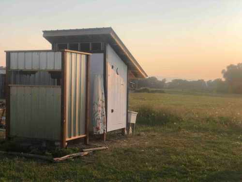 A small shed with a sloped roof stands in a grassy field at sunrise, surrounded by a peaceful rural landscape.