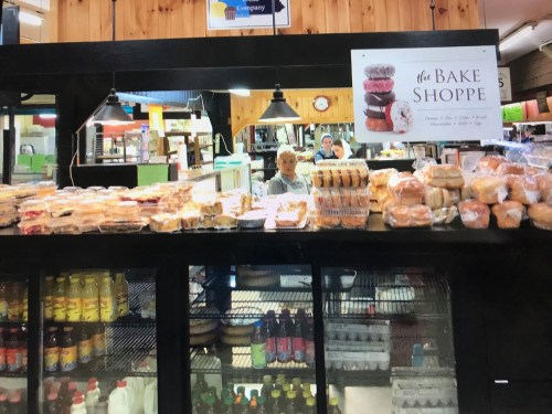 A bakery display filled with various baked goods, with a sign reading "The Bake Shoppe" and a staff member behind the counter.