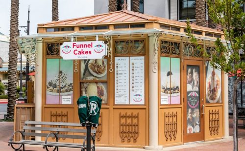 A small kiosk selling funnel cakes, featuring a menu and outdoor seating, surrounded by palm trees.