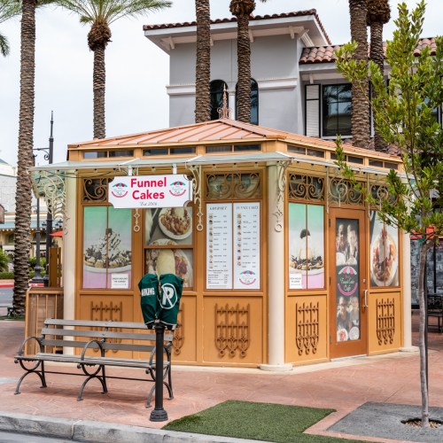 A colorful funnel cake stand with a menu, surrounded by palm trees and a bench in a lively outdoor setting.