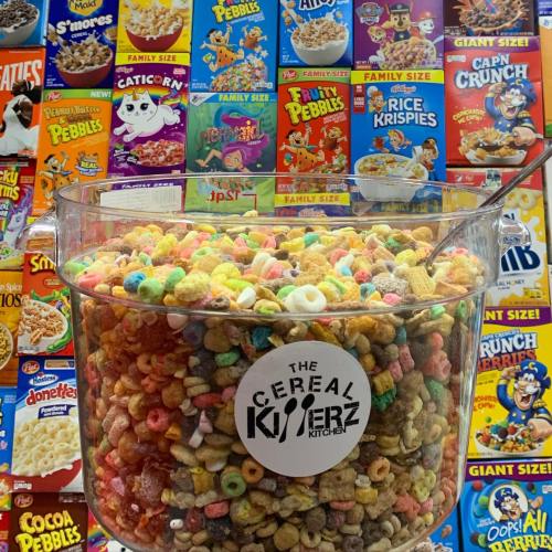 A large container filled with colorful cereal, surrounded by various cereal boxes on a wall.