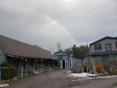 A quaint building with a sloped roof and a rainbow in the sky, surrounded by trees and a snowy path.