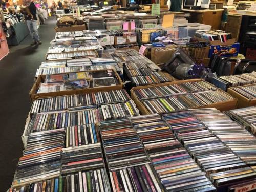 A large collection of CDs displayed on tables in a bustling market, with shoppers browsing in the background.