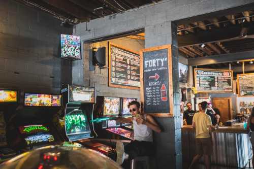 A retro arcade with colorful game machines, a man in sunglasses relaxes by the wall, and a bar area is visible in the background.