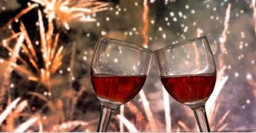 Two glasses of red wine clink together against a backdrop of fireworks and sparkling lights.