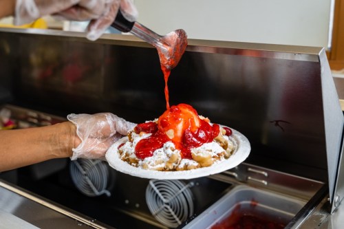 A hand in a glove drizzles strawberry sauce over a plate of funnel cake topped with whipped cream and strawberries.