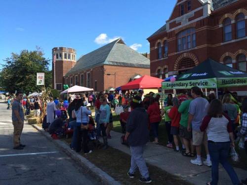 A lively street fair with people enjoying food and activities near tents and a historic building under a clear blue sky.
