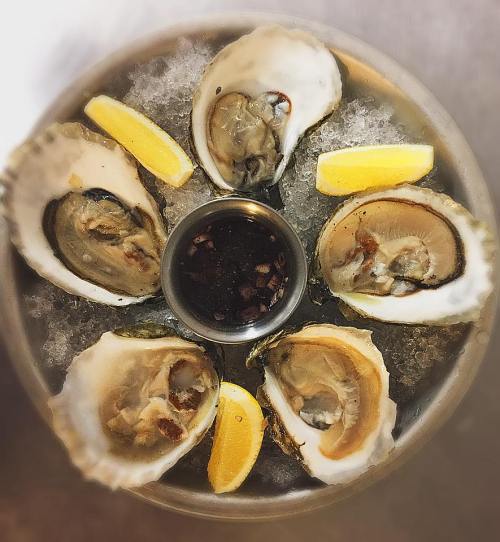 A platter of fresh oysters on ice, garnished with lemon wedges and a small bowl of dipping sauce.