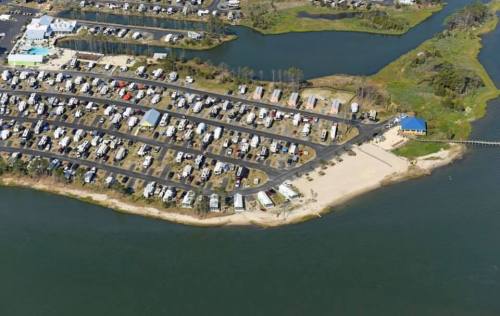 Aerial view of a campground with RVs, a beach area, and a winding river nearby. Green spaces and water surround the site.