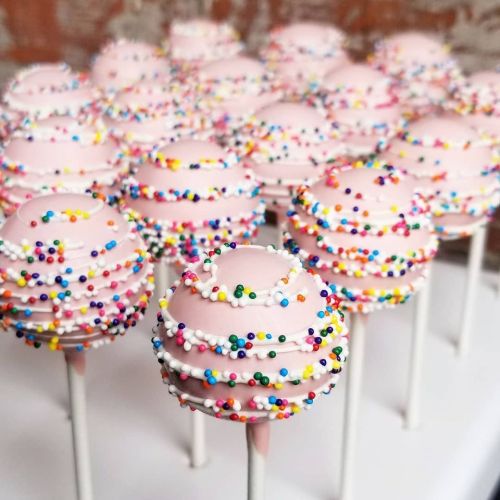 A close-up of pink cake pops decorated with colorful sprinkles on white sticks, arranged in a neat display.