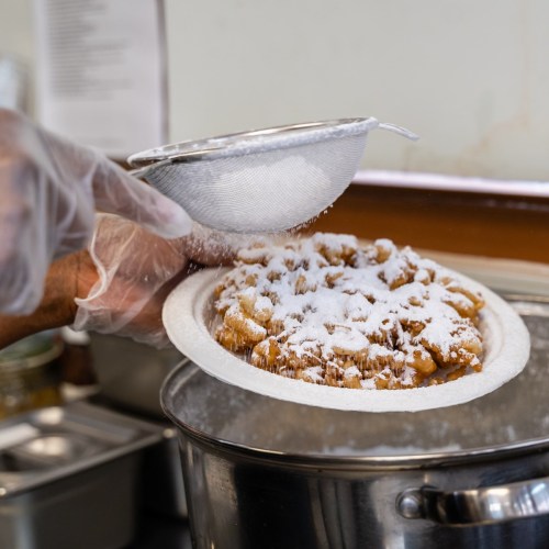 A hand sifts powdered sugar over a freshly baked dessert on a plate, with kitchen items in the background.