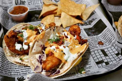 Two tacos filled with fried fish, slaw, and sauce, served on newspaper with tortilla chips and a small cup of salsa.