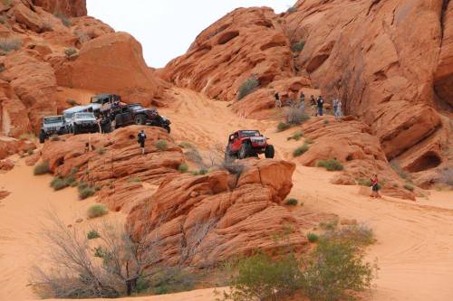 Off-road vehicles navigate rocky terrain in a desert landscape, with people observing from the rocks above.