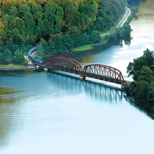 Aerial view of a red bridge over a river, surrounded by lush green trees and a winding road along the water's edge.