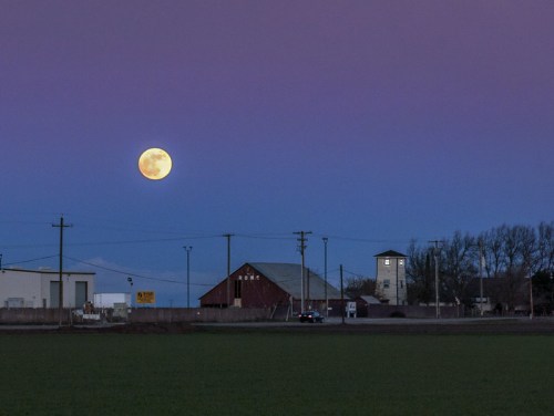 A full moon rises over a rural landscape with a red barn and a water tower against a twilight sky.